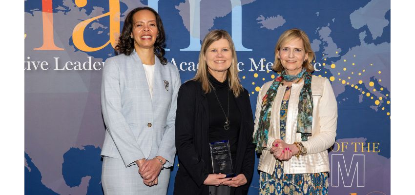 Three women stand in front of a backdrop at a professional event, one holding an award plaque.
