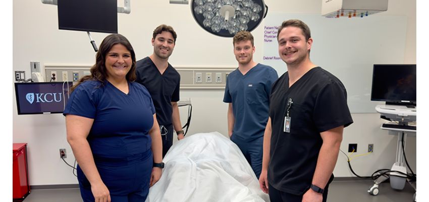 Four medical students in scrubs pose in a clinical simulation room.