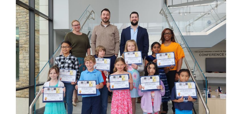 A group of elementary students proudly hold up certificates while standing on stairs with adults behind them at a school or community event.