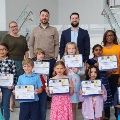Agroupofelementarystudentsproudlyholdupcertificateswhilestandingonstairswithadultsbehindthemataschoolorcommunityevent