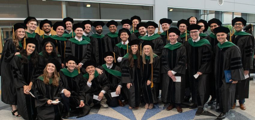A large group of graduating medical students in academic regalia pose together indoors, smiling at the camera.
