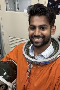 A man smiles while wearing an orange astronaut training suit, holding a helmet.