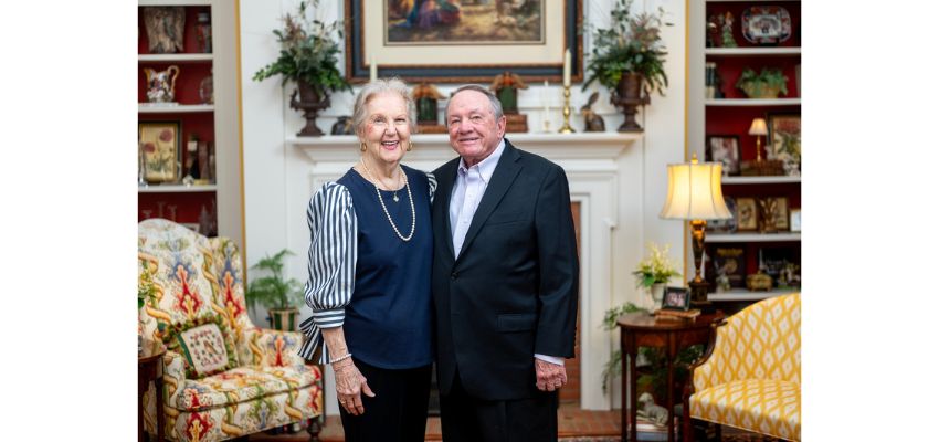 An older couple stands side by side and smiles in a warmly decorated living room with bookshelves and traditional furniture.