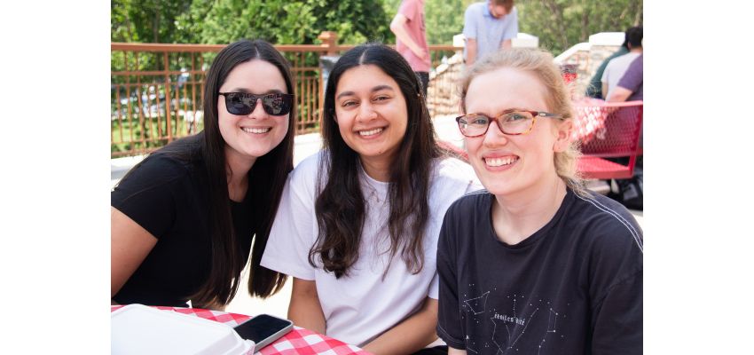 Three young women sit at an outdoor picnic table with checkered cloth, smiling on a sunny day.