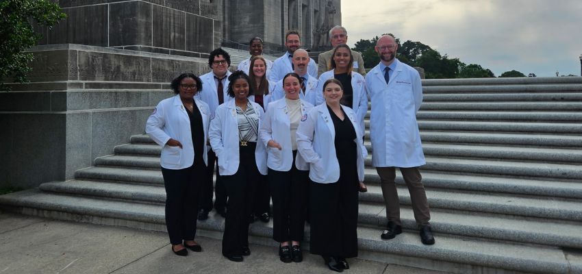 A group of students and faculty in white coats pose together on the steps of a large stone building, likely a government or historic site.