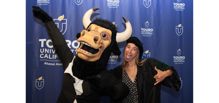 A Touro University California graduate poses joyfully with the university’s mascot in front of a branded backdrop.