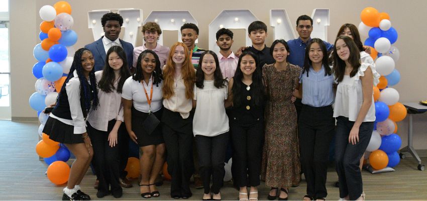 A diverse group of students dressed in business casual clothing pose together under a balloon arch with VCOM letters behind them.