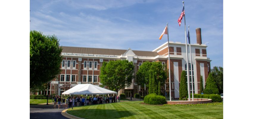 People gather under a white event tent on the lawn of a red-brick medical school building with flags flying in the foreground.