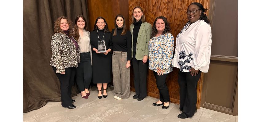 A group of women pose together indoors, one holding an award, smiling in celebration of a professional recognition.