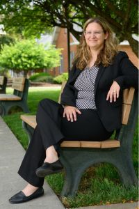 A woman in a black suit sits confidently on a bench outside, smiling and relaxed on a sunny day.