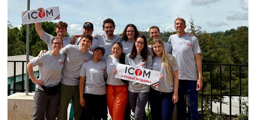 A group of ICOM medical students pose outdoors holding “ICOM Global Brigades” signs during an international service trip.