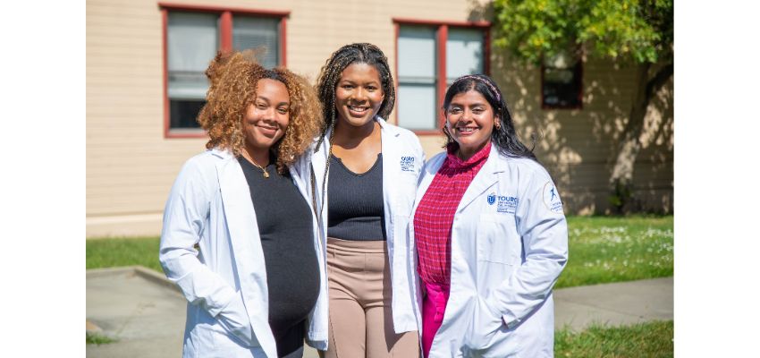 Three Touro University California medical students in white coats stand smiling together outside on campus.