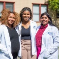 ThreeTouroUniversityCaliforniamedicalstudentsinwhitecoatsstandsmilingtogetheroutsideoncampus