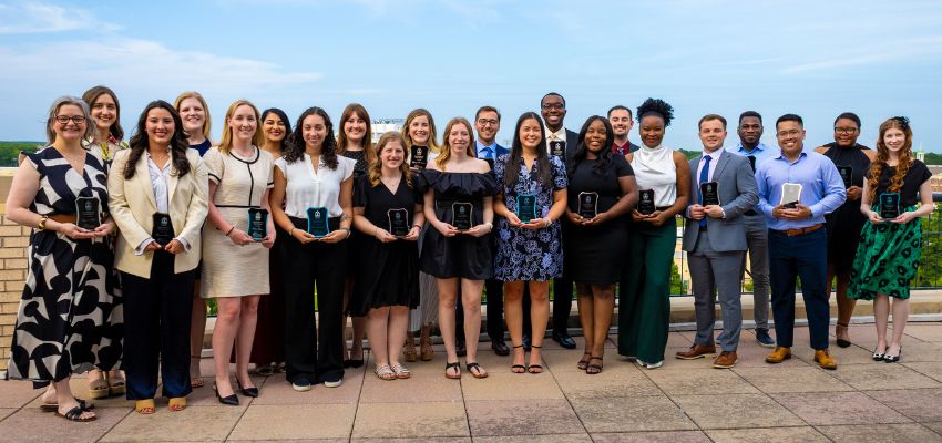 A group of VCOM-Louisiana students and faculty pose with awards on a rooftop terrace overlooking a scenic backdrop.