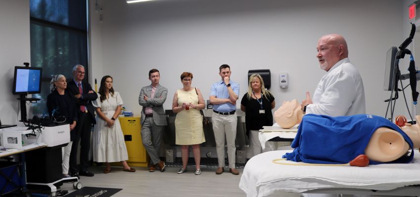 A medical educator speaks to a group gathered around a simulation lab, demonstrating with a medical mannequin.