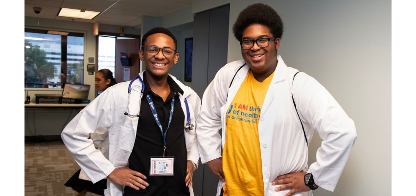 Two medical students wearing white coats smile in a clinical setting, showing enthusiasm for their training.