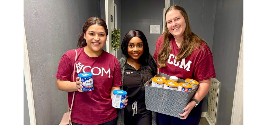 Three individuals, two wearing VCOM shirts, hold baby formula donations while standing together in a community service setting.