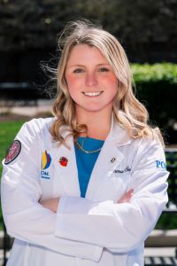 A PCOM student wearing a white coat smiles confidently outdoors with arms crossed.