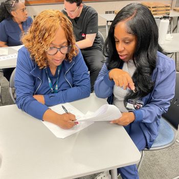 Two women in medical scrubs collaborate while reviewing paperwork during a classroom training session.