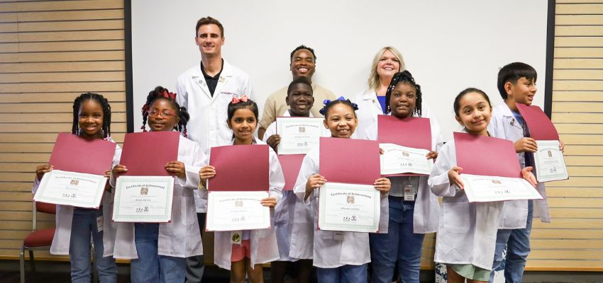 Elementary school students proudly hold up certificates while wearing white coats alongside VCOM-Louisiana mentors.