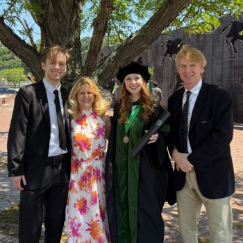 A newly graduated medical student in cap and gown poses with her family under a tree on a sunny day.