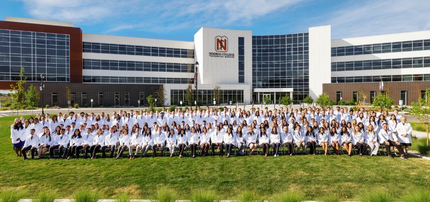 A large group of medical students in white coats pose for a class photo in front of Noorda College of Osteopathic Medicine.