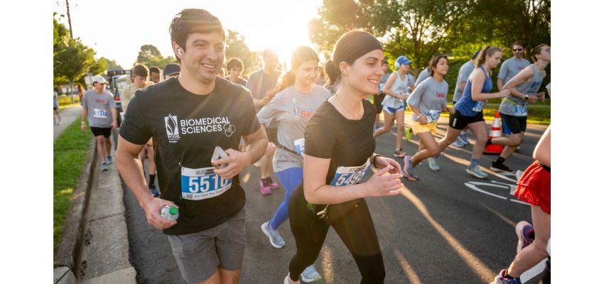 Participants, including a man wearing a PCOM Georgia Biomedical Sciences shirt, run in a 5K race during daylight.