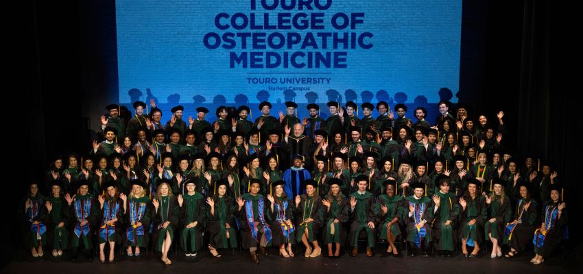 A large group of graduates in green academic regalia pose on stage in front of a screen reading “Touro College of Osteopathic Medicine – Touro University Harlem Campus.”