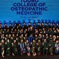 AlargegroupofgraduatesingreenacademicregaliaposeonstageinfrontofascreenreadingTouroCollegeofOsteopathicMedicineTouroUniversityHarlemCampus