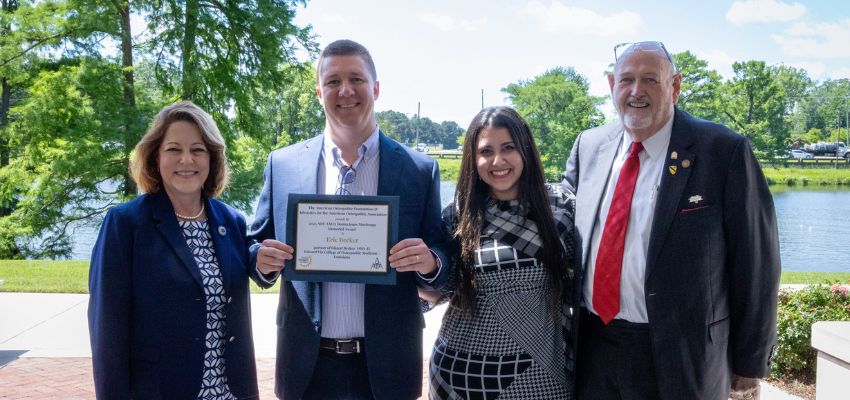 A man holds a framed award certificate while posing with three smiling adults near a lakeside on a sunny day.
