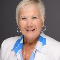 Awomanwithshortwhitehairinawhiteblazerandblueshirtsmilesinaprofessionalheadshotagainstagraybackground