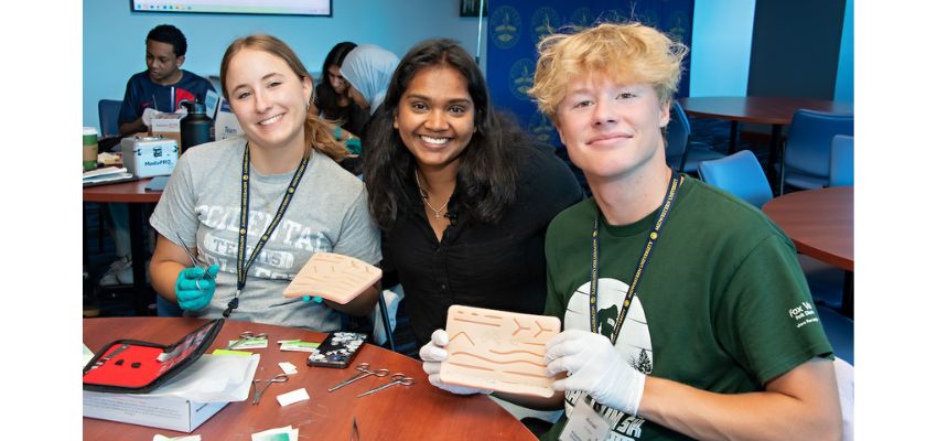 Three students in a classroom setting smile and hold up suture practice pads during a hands-on medical training activity.