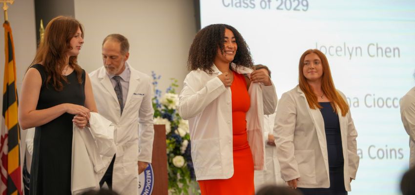 A student in a red dress puts on a white coat during a white coat ceremony while others on stage look on.