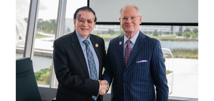 Two professionally dressed men shake hands and smile in front of a window with a lake and buildings in the background.