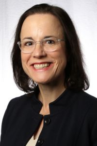 Headshot of a smiling woman in glasses and a dark blazer in front of a white backdrop.