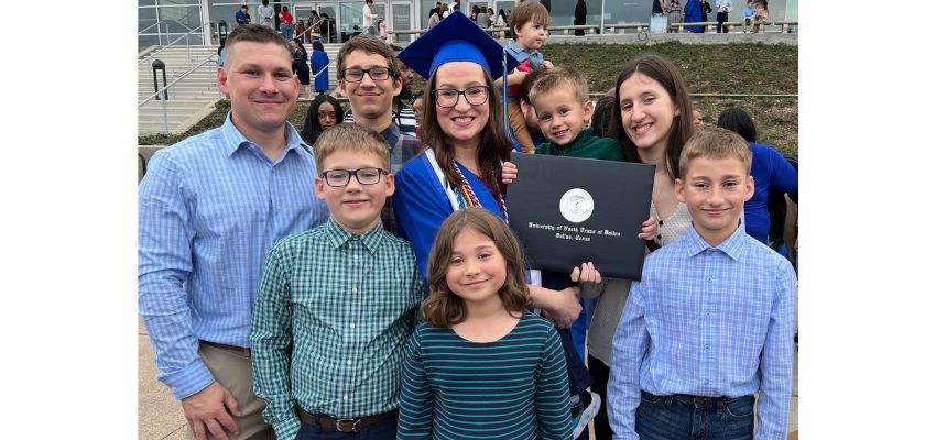 A graduate in a blue cap and gown holds a diploma while surrounded by smiling family members outside the ceremony venue.