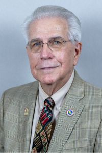 Portrait of an older man in a plaid suit jacket and patterned tie, smiling at the camera against a gray background.