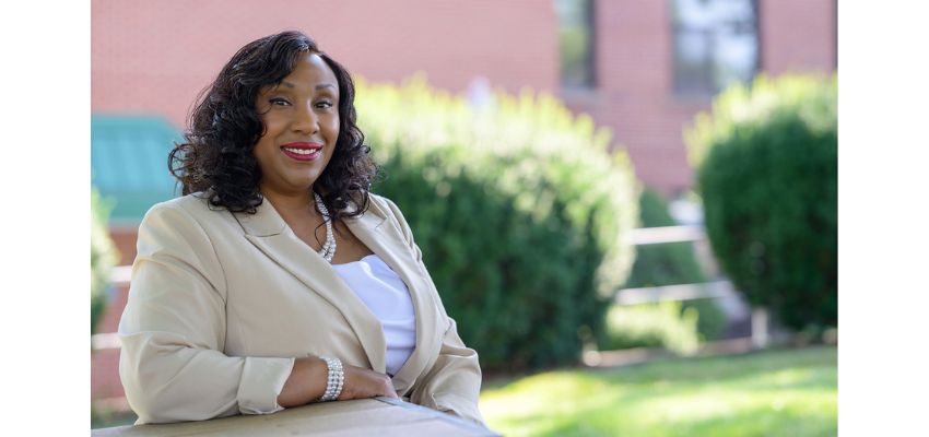 A woman in a beige suit jacket smiles while seated outdoors in front of greenery and a brick building.