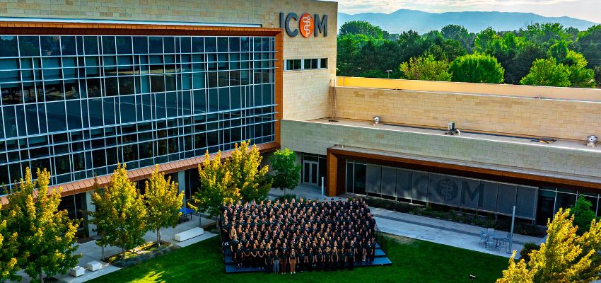 A large group of students pose for a photo outside the Idaho College of Osteopathic Medicine with trees and mountains in the background.