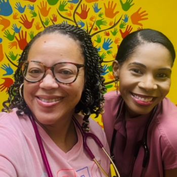 Two women wearing stethoscopes smile in front of a colorful mural featuring a tree made of multicolored handprints.