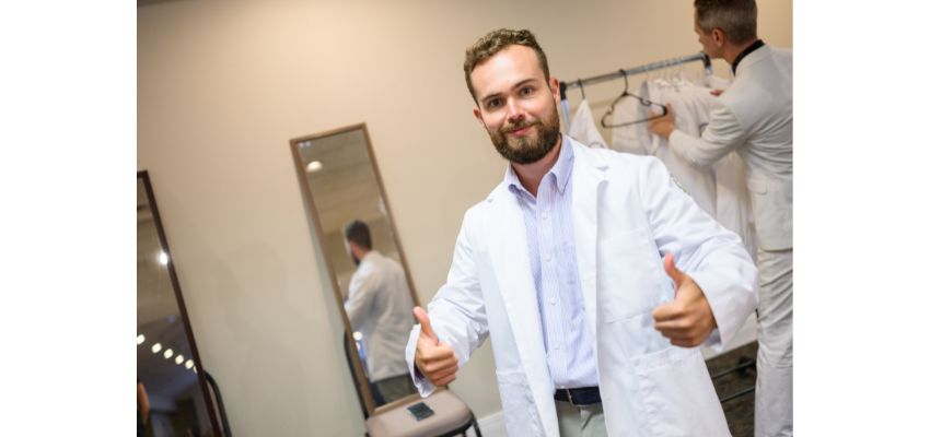A man in a white coat gives two thumbs up and smiles at the camera while another person arranges coats in the background.