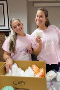 Two women wearing matching pink shirts smile while standing next to a box filled with feminine hygiene products.