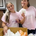 Twowomenwearingmatchingpinkshirtssmilewhilestandingnexttoaboxfilledwithfemininehygieneproducts