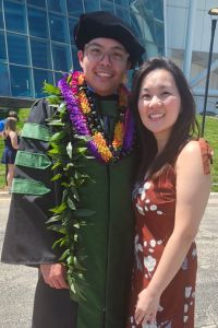 A graduate in a green cap and gown with flower leis around his neck stands smiling next his wife outdoors.