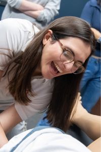 A woman wearing glasses and a white shirt laughs while leaning over during a hands-on activity with classmates.