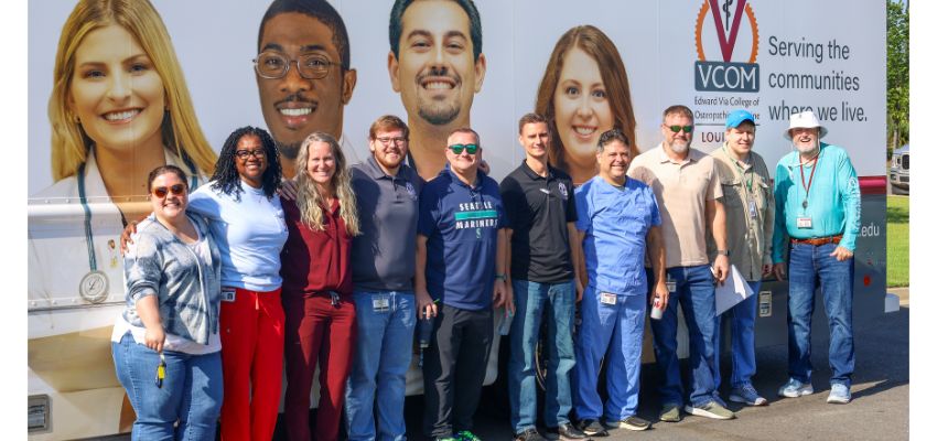 A group of people stand smiling in front of a VCOM Louisiana mobile clinic vehicle with portraits of students printed on the side.