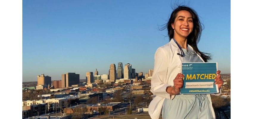 A smiling medical student in a white coat holds a “Matched” sign in front of a city skyline.