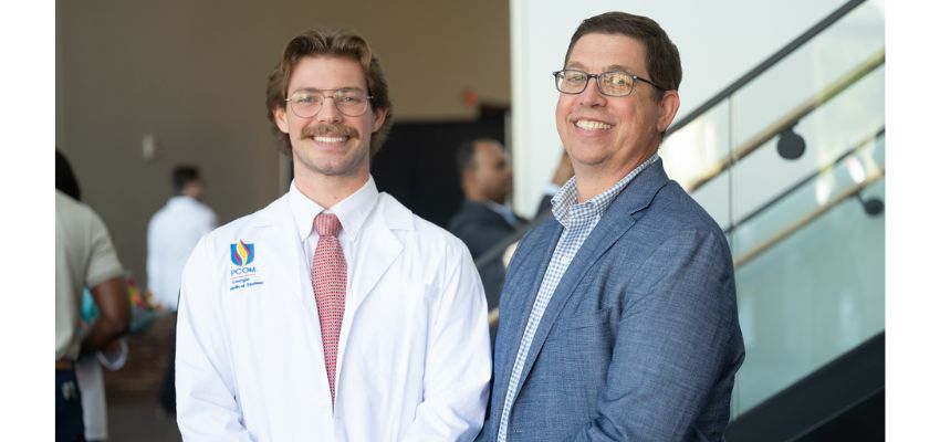 A student in a white coat stands beside a man in a suit, both smiling at the camera in a bright indoor space.