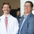 Astudentinawhitecoatstandsbesideamaninasuitbothsmilingatthecamerainabrightindoorspace