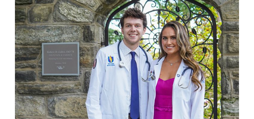 Two medical students wearing white coats and stethoscopes smile while standing together outside near a stone archway.